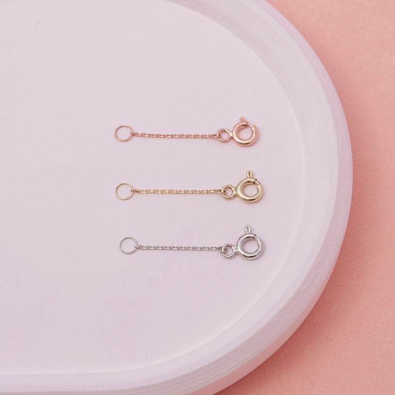May include: Three necklace extenders in rose gold, gold, and silver. Each extender features a small chain with a spring ring clasp on one end and a small ring on the other. The extenders are displayed on a white, curved surface against a pink background.