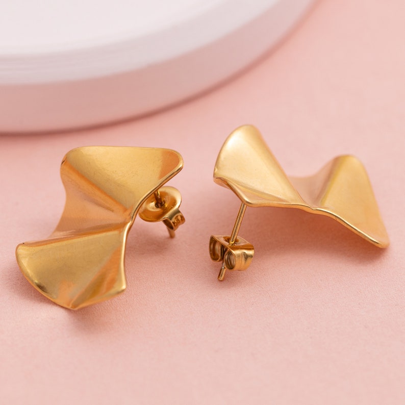 May include: A pair of gold-toned earrings with a wavy, asymmetrical design. The earrings are made of metal and have a post-back closure.