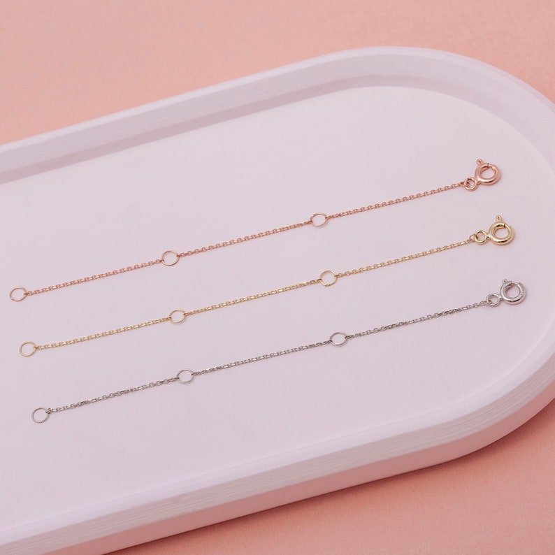 May include: Three delicate chain extenders in rose gold, gold, and silver. Each extender features a small chain with a clasp on one end and a series of jump rings for adjustable length. The extenders are displayed on a white tray against a pink background.