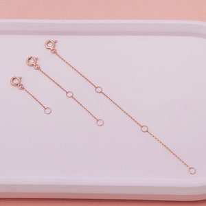 May include: Three rose gold necklace extenders of varying lengths. Each extender features a lobster clasp on one end and a small ring on the other. The extenders also have small rings along the chain for length adjustment. The items are displayed on a white tray.