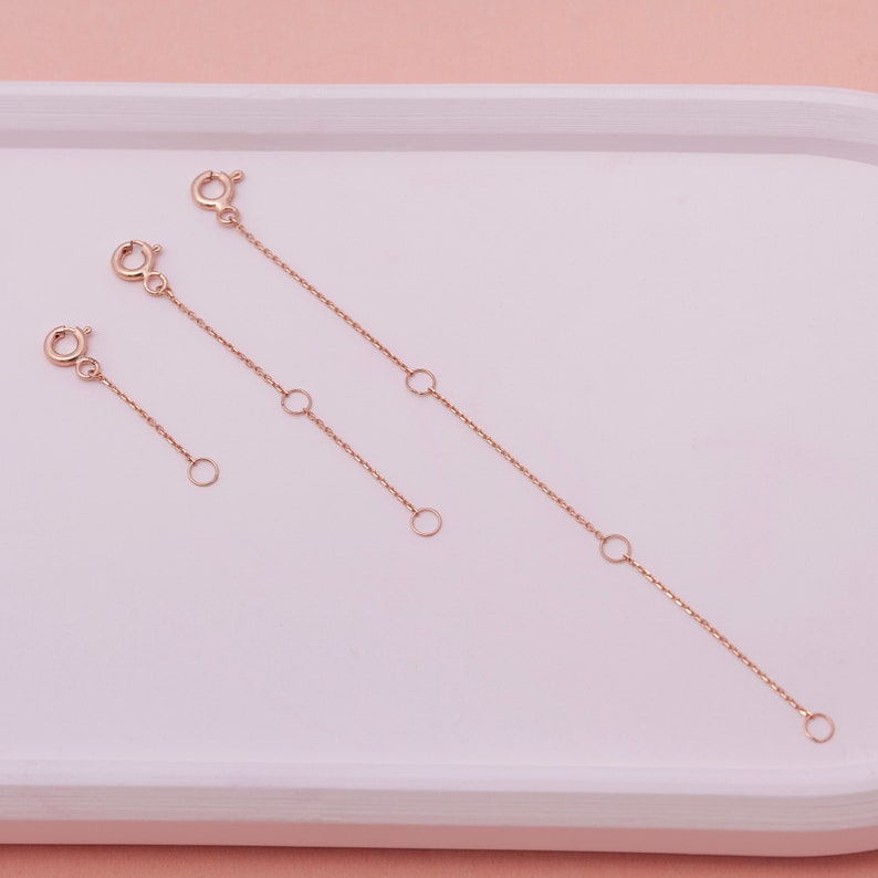 May include: Three rose gold necklace extenders of varying lengths. Each extender features a lobster clasp on one end and a small ring on the other. The extenders also have small rings along the chain for length adjustment. The items are displayed on a white tray.