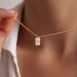 May include: A gold chain necklace with a small rectangular pendant featuring the letter 'E' and a tiny diamond.