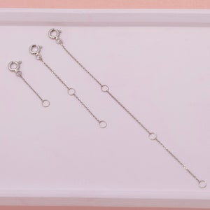 May include: Three silver-colored chain extenders of varying lengths. Each extender features a lobster clasp on one end and a small ring on the other. The chains have small, circular accent rings along their length. The extenders are displayed on a light pink surface.