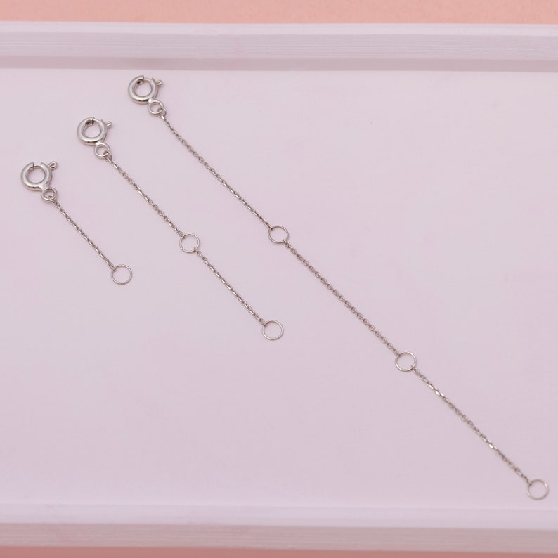 May include: Three silver-colored chain extenders of varying lengths. Each extender features a lobster clasp on one end and a small ring on the other. The chains have small, circular accent rings along their length. The extenders are displayed on a light pink surface.