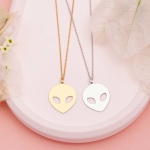 Pu&ograve; includere: Due delicate collane con ciondoli a forma di testa aliena. Una collana &egrave; dorata e l'altra &egrave; argentata. Le collane sono esposte su una superficie bianca.