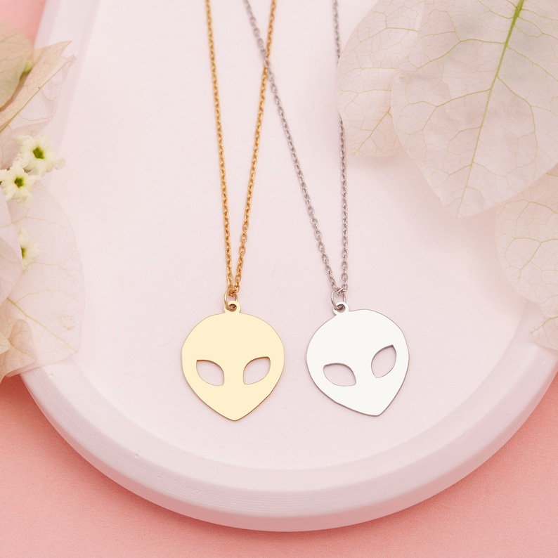 Pu&ograve; includere: Due delicate collane con ciondoli a forma di testa aliena. Una collana &egrave; dorata e l'altra &egrave; argentata. Le collane sono esposte su una superficie bianca.