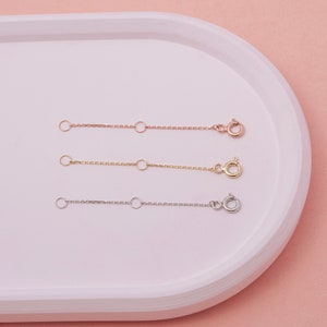 May include: Three necklace extender chains in rose gold, gold, and silver. Each chain has a small clasp on one end and a series of small rings for adjusting the length. The chains are displayed on a white tray against a pink background.