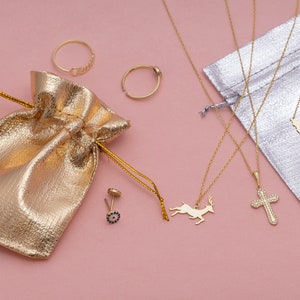 May include: A gold pouch with a variety of gold jewelry on a pink background. The jewelry includes a ring, a necklace with a deer pendant, a necklace with a cross pendant, a necklace with a small island pendant, and a pair of earrings with blue stones.