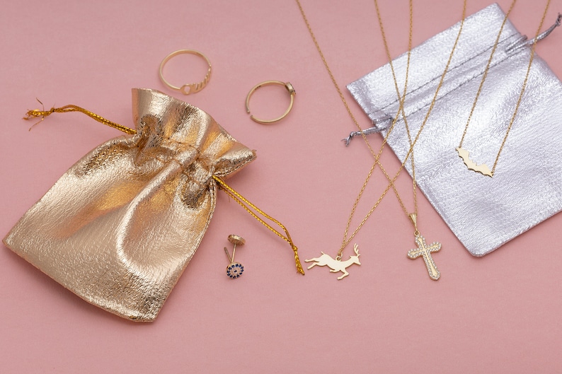 May include: A gold pouch with a variety of gold jewelry on a pink background. The jewelry includes a ring, a necklace with a deer pendant, a necklace with a cross pendant, a necklace with a small island pendant, and a pair of earrings with blue stones.