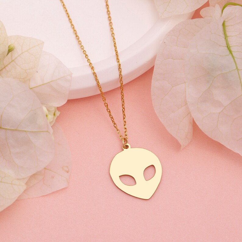 Pu&ograve; includere: Una collana con catena dorata con un ciondolo a forma di testa aliena. Il ciondolo &egrave; un semplice contorno di una testa aliena con occhi grandi.