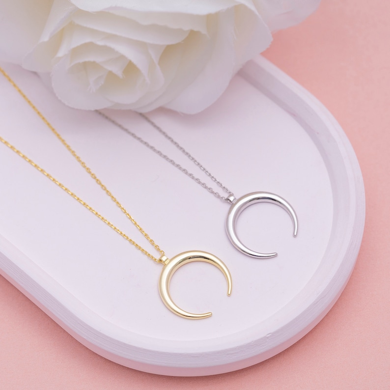 May include: Two delicate necklaces with crescent moon pendants. One necklace is gold and the other is silver. The necklaces are displayed on a white oval dish.