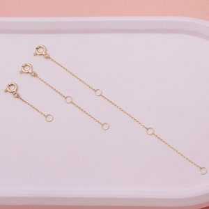 May include: Three gold-colored chain extenders of varying lengths. Each extender features a clasp on one end and a small ring on the other, with additional rings spaced along the chain. The extenders are designed to add length to necklaces or bracelets.