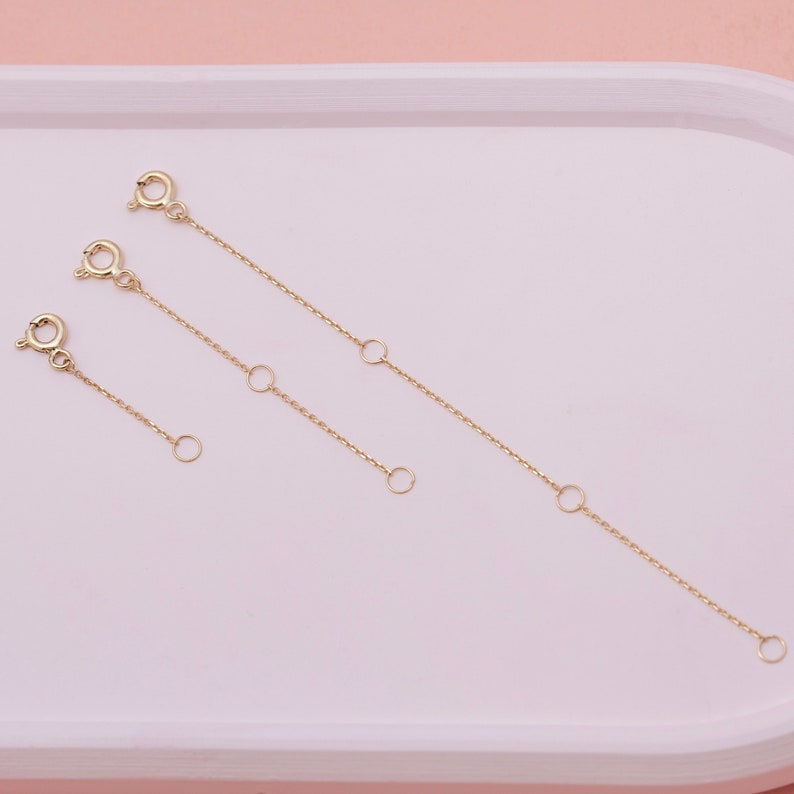 May include: Three gold-colored chain extenders of varying lengths. Each extender features a clasp on one end and a small ring on the other, with additional rings spaced along the chain. The extenders are designed to add length to necklaces or bracelets.