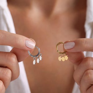 May include: Two hoop earrings with dangling charms. The earring on the left is silver with three silver charms. The earring on the right is gold with three gold charms.