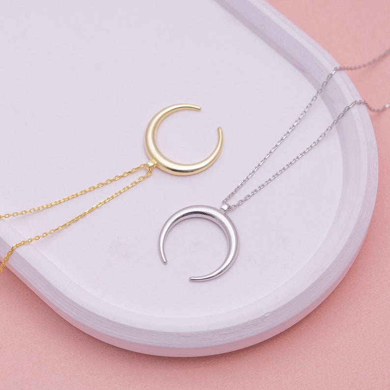 May include: Two delicate crescent moon necklaces, one in gold and one in silver, displayed on a white surface. The necklaces feature a simple chain design.