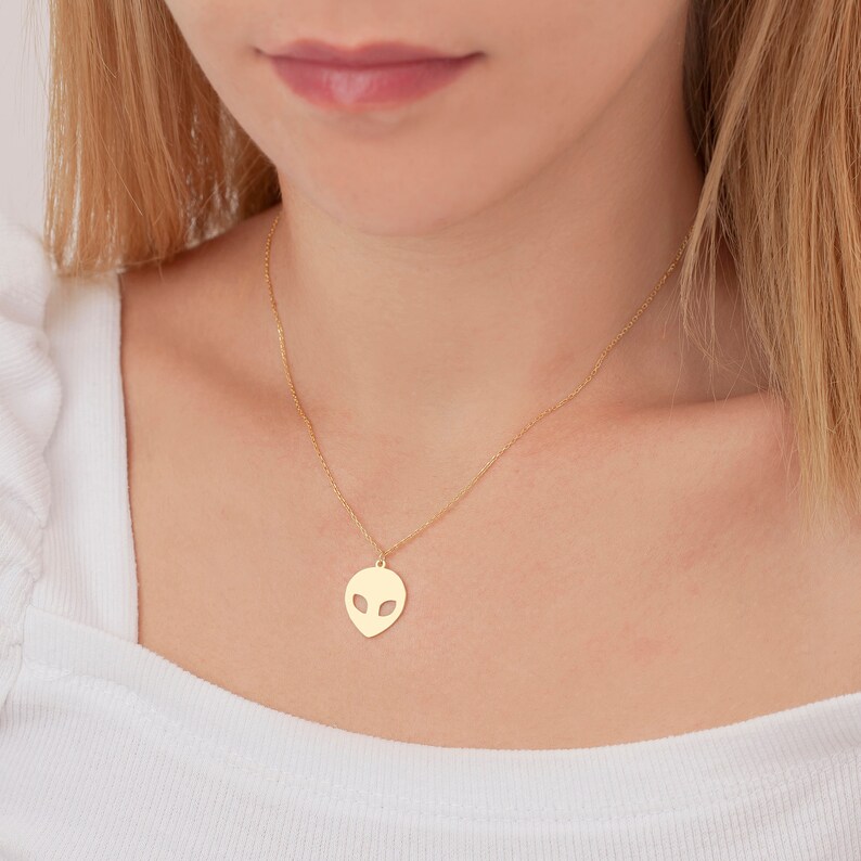 Pu&ograve; includere: Una collana con catena dorata con un piccolo ciondolo a forma di testa aliena. Il ciondolo &egrave; un semplice contorno di un viso alieno con occhi grandi.