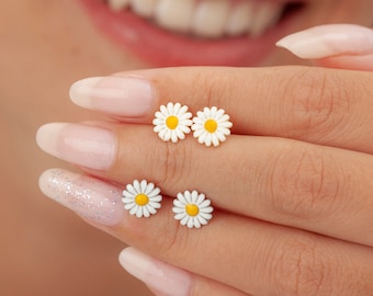 May include: Four white daisy earrings with yellow centers. The earrings are on a hand with manicured nails.