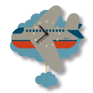 May include: A wooden airplane-shaped wall clock with a light gray body, orange and blue accents, and black clock hands. The clock is surrounded by a blue cloud design and includes the text "popclox". The clock measures approximately 12 inches in length.
