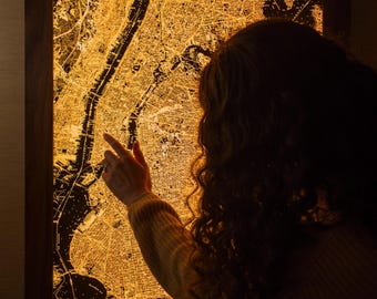 Peut inclure: Une carte illuminée de New York encadrée, brillant d'une lumière dorée et chaleureuse. La carte est détaillée, montrant les rues et les cours d'eau. Le cadre est en bois foncé. Une main pointe la carte.