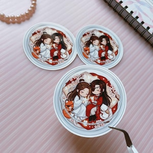May include: Three circular stickers featuring a chibi-style illustration of two characters with long dark hair, one in white robes and one in red robes, sitting together surrounded by cherry blossoms, a small orange fox, and a white cat.