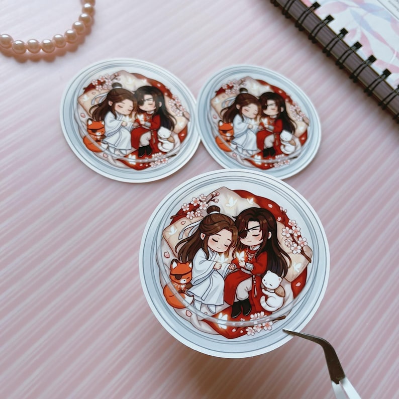 May include: Three circular stickers featuring a chibi-style illustration of two characters with long dark hair, one in white robes and one in red robes, sitting together surrounded by cherry blossoms, a small orange fox, and a white cat.