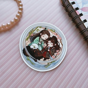 May include: A circular sticker featuring a chibi-style illustration of two figures with long dark hair, one in green and one in dark red robes, sleeping together. A small white cat and a black dog rest beside them, surrounded by bamboo and pink blossoms.