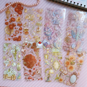 May include: A sheet of clear, transparent stickers with a variety of floral and vintage-inspired designs. The stickers feature images of roses, lilies, sunflowers, birds, cages, books, and other decorative elements. The stickers are perfect for adding a touch of whimsy and charm to journals, planners, and other craft projects.