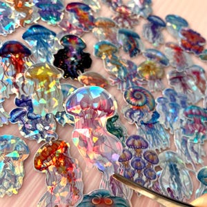 May include: A collection of holographic jellyfish stickers featuring various colours including blue, purple, red, and yellow. Each sticker displays a unique jellyfish shape with a reflective, prismatic finish that catches the light, creating a shimmering effect across the surface of the designs.