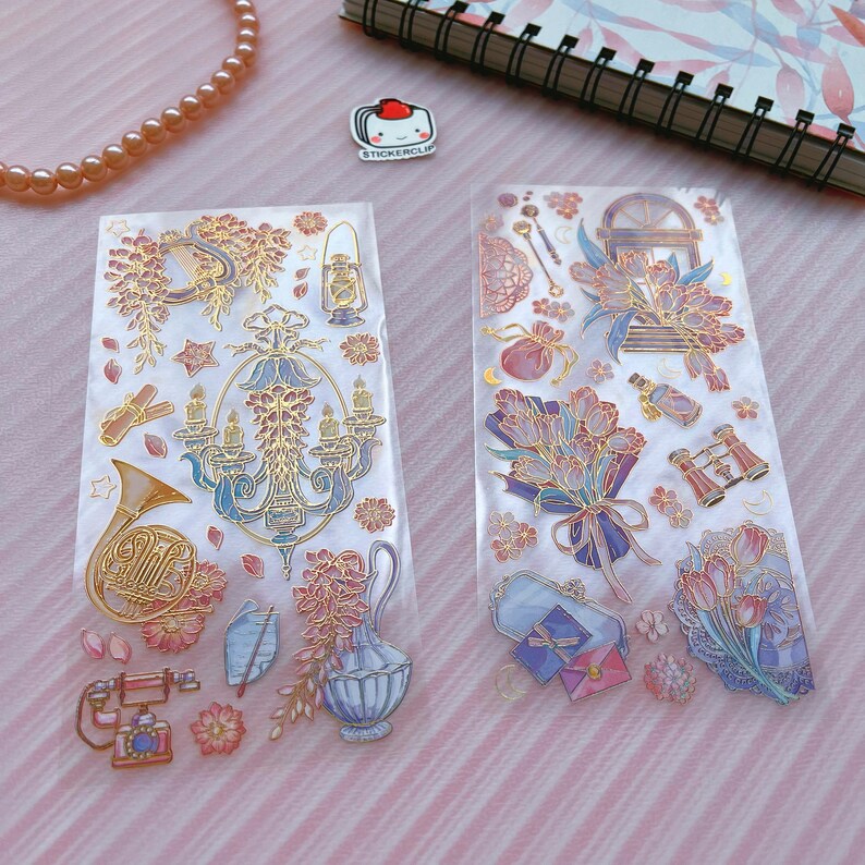 May include: Two sheets of clear sticker paper with gold and pastel colored illustrations. The first sheet features a gold french horn, a chandelier, a lantern, and floral designs. The second sheet features a bouquet of flowers, a pair of binoculars, a purse, and a window.