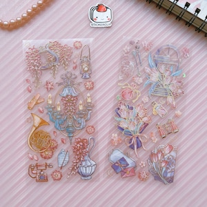 May include: Two sheets of clear sticker paper with gold and pink floral designs. The stickers feature a chandelier, a bouquet of tulips, a vintage-style camera, a french horn, a lantern, a bottle, and a pair of binoculars.