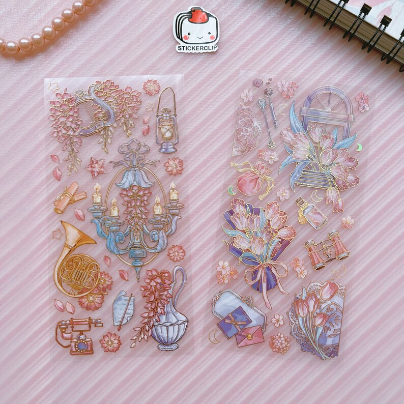 May include: Two sheets of clear sticker paper with gold and pink floral designs. The stickers feature a chandelier, a bouquet of tulips, a vintage-style camera, a french horn, a lantern, a bottle, and a pair of binoculars.