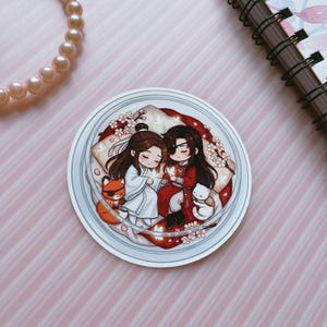 May include: A circular sticker featuring a chibi-style illustration of two figures with long dark hair, one in white and one in red robes, resting on pillows. They are surrounded by small white flowers, a small orange fox, and a white cat-like creature.