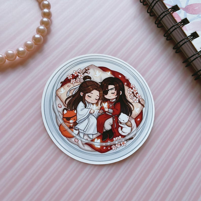 May include: A circular sticker featuring a chibi-style illustration of two figures with long dark hair, one in white and one in red robes, resting on pillows. They are surrounded by small white flowers, a small orange fox, and a white cat-like creature.