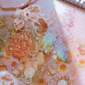 May include: A sheet of gold foil stickers with various designs, including roses, sunflowers, bottles, and a birdcage. The stickers are on a pink background.