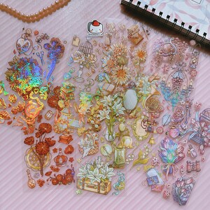 May include: A sheet of iridescent holographic stickers featuring a variety of designs, including flowers, books, musical instruments, and other whimsical elements. The stickers are perfect for adding a touch of sparkle and magic to journals, planners, and other craft projects.