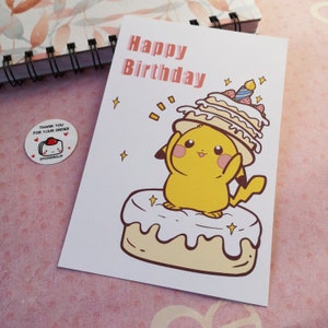 May include: A greeting card with a yellow cartoon character wearing a cake hat. The card says "Happy Birthday" in pink letters. The character is standing on a cake with white frosting.