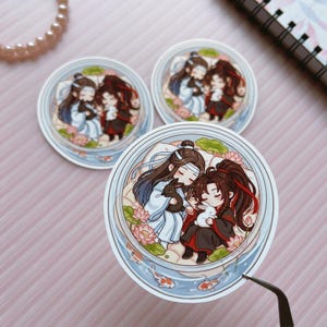 May include: Three circular stickers featuring a chibi-style illustration of two characters with long hair, one in white and blue robes and one in black and red robes, each holding a small rabbit amidst pink lotus flowers and koi fish in water.