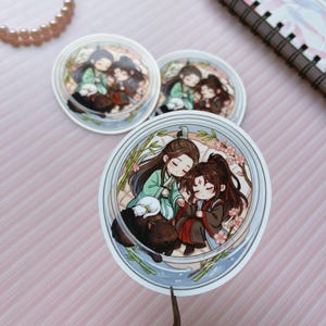 May include: Three circular stickers featuring a chibi-style illustration of two characters with long dark hair, one in green and one in red robes, resting together. A small white cat and a black dog are nearby, surrounded by bamboo and pink blossoms.