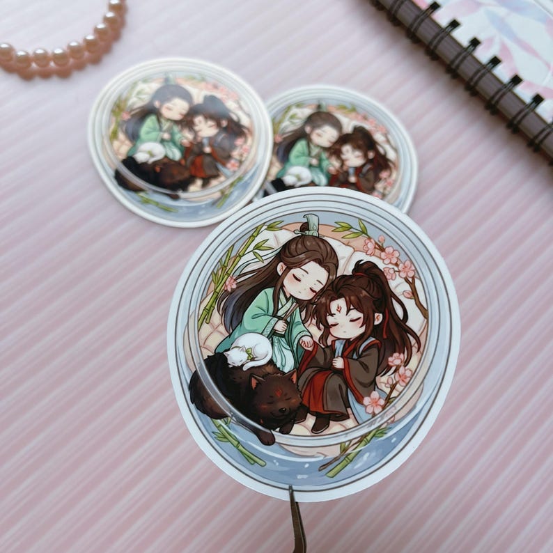 May include: Three circular stickers featuring a chibi-style illustration of two characters with long dark hair, one in green and one in red robes, resting together. A small white cat and a black dog are nearby, surrounded by bamboo and pink blossoms.