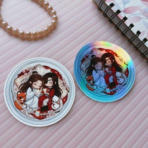 May include: Two circular stickers featuring a chibi-style illustration of two characters with long dark hair, one in white and one in red robes, resting together amidst cherry blossoms, a small fox, and a white cat. One sticker has a holographic finish.