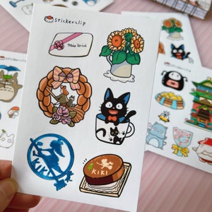 May include: A sheet of stickers featuring characters from the animated film Kiki's Delivery Service. The stickers include Kiki the witch, Jiji the black cat, a sunflower bouquet, a wreath, and a cake. The stickers are printed on white paper with a glossy finish.