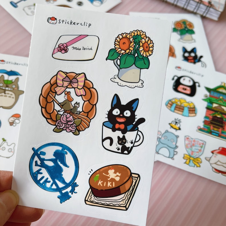 May include: A sheet of stickers featuring characters from the animated film Kiki's Delivery Service. The stickers include Kiki the witch, Jiji the black cat, a sunflower bouquet, a wreath, and a cake. The stickers are printed on white paper with a glossy finish.