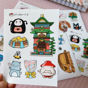 May include: A sheet of stickers featuring characters and scenes from the popular animated film Spirited Away. The stickers include a red and green building, a white and blue bathhouse, a white and blue cat-like creature, a red and white creature with a red hat, a white and blue bird, and a brown and white creature with large ears. The sheet also includes the text "Stickerclip".