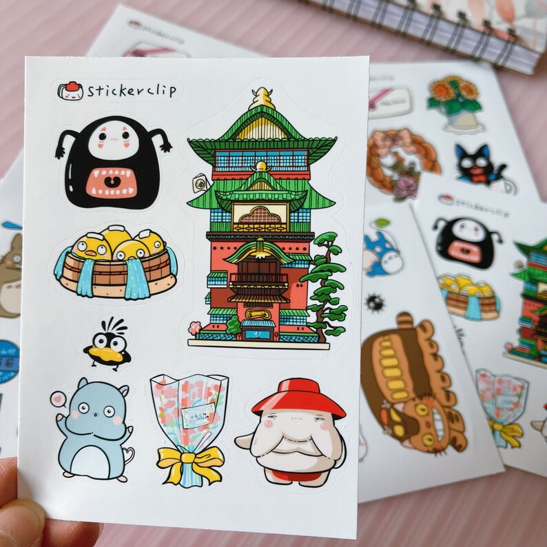 May include: A sheet of stickers featuring characters and scenes from the popular animated film Spirited Away. The stickers include a red and green building, a white and blue bathhouse, a white and blue cat-like creature, a red and white creature with a red hat, a white and blue bird, and a brown and white creature with large ears. The sheet also includes the text "Stickerclip".