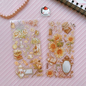 May include: Two sheets of clear sticker paper with gold foil accents. The first sheet features a floral arrangement with a birdcage, a lamp, a teapot, books, and other decorative elements. The second sheet features sunflowers, a mirror, books, and other decorative elements.