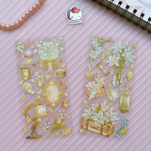 May include: Two sheets of clear sticker paper with gold foil accents. The stickers feature illustrations of lilies, bells, a watering can, a mirror, a lantern, a suitcase, and other vintage-inspired designs.