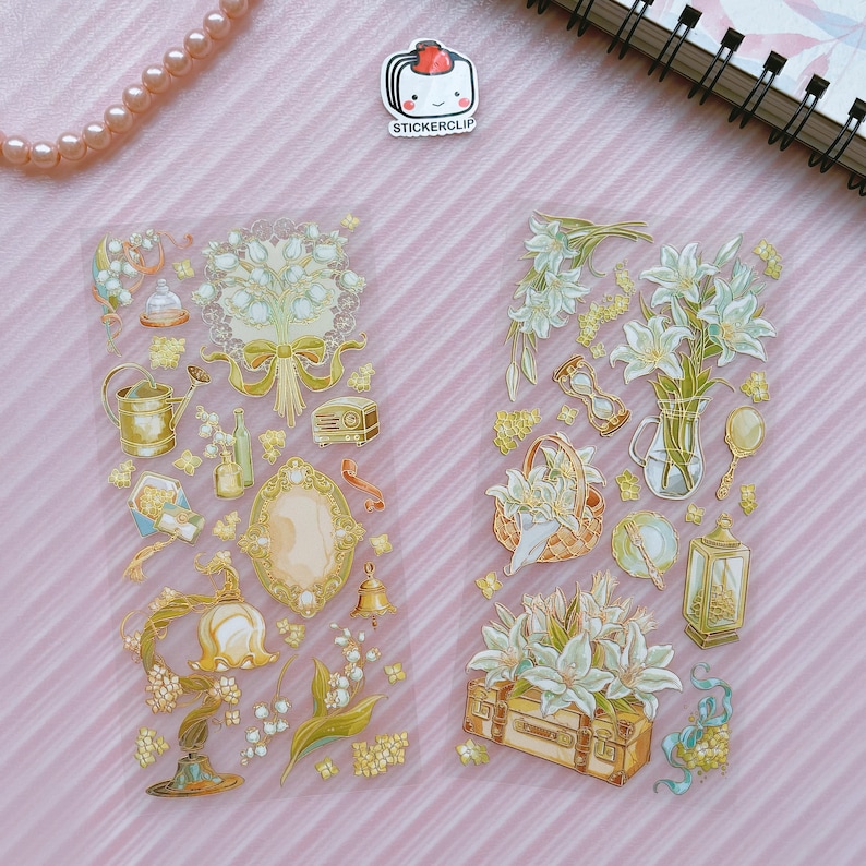 May include: Two sheets of clear sticker paper with gold foil accents. The stickers feature illustrations of lilies, bells, a watering can, a mirror, a lantern, a suitcase, and other vintage-inspired designs.
