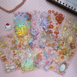 May include: A sheet of iridescent holographic stickers featuring floral bouquets, a window, a birdcage, a teapot, a mirror, a french horn, and other decorative elements. The stickers are on a pink background.