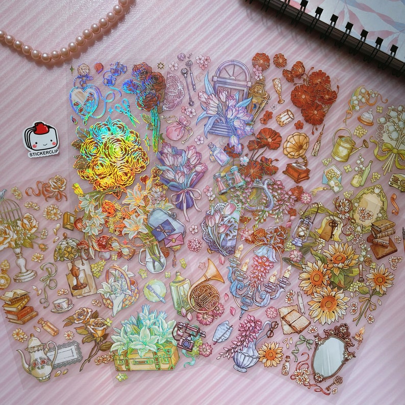 May include: A sheet of iridescent holographic stickers featuring floral bouquets, a window, a birdcage, a teapot, a mirror, a french horn, and other decorative elements. The stickers are on a pink background.