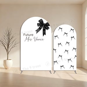 May include: Two white arched backdrop stands. One features a large black bow and the text "Future Mrs Vance." The other has a pattern of small black bows. Both stands have metal supports and are for a wedding or bridal event.